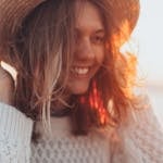 Joyful woman in a straw hat smiling by the seaside with sunset glow.